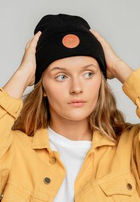 Czarna dziana czapka beanie z okrągłym, pomarańczowym logo przedstawiającym górę i zwierzę. Noszona z żółtą kurtką i białą koszulą pod spodem.