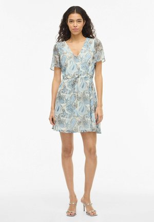 Moteris su ilgais garbanotais plaukais, dėvinti šviesiai mėlyną paisley raštuotą suknelę su raišteliais ir sidabrines aukštakulnes basutes, stovinti prie balto fono.