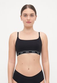 Bralette nera con scollatura quadrata, spalline sottili e un elastico con marchio che presenta lettere bianche. Tessuto liscio, design minimale.
