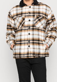 Man i en knapplös fleecejacka med svart, vit och brun rutmönster och svarta byxor, händerna i fickorna.