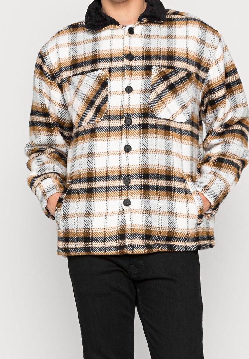 Man i en knapplös fleecejacka med svart, vit och brun rutmönster och svarta byxor, händerna i fickorna.