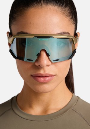 Donna che indossa grandi occhiali sportivi con lenti scure, montatura dorata e naselli neri, vestita con una maglia verde oliva.