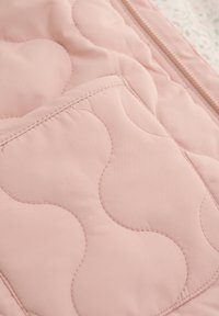Quiltad rosa tyg med ett vågigt mönster, med en synlig ficka och metallblixtlås. Mjuk textur, slät yta och diskret design.