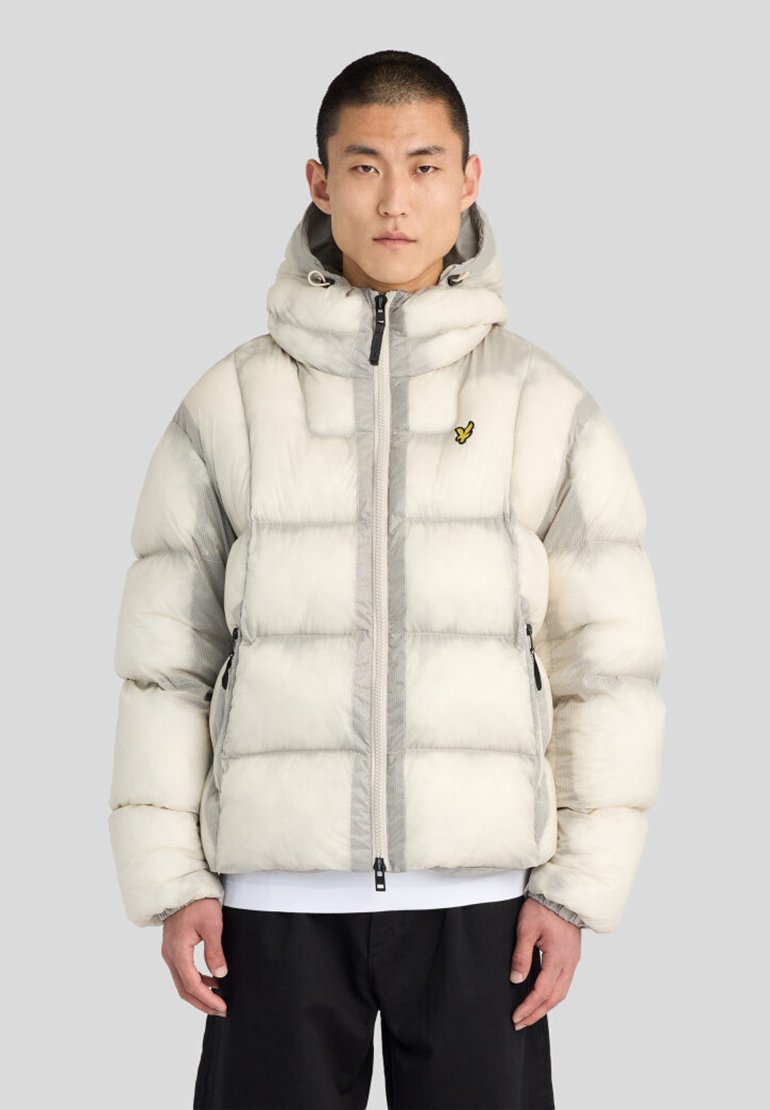 Jeune homme portant une veste matelassée à capuche gris clair avec un petit logo d’oiseau jaune sur la poitrine et un pantalon noir.