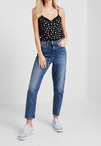 Camisole nera con stampa floreale e spalline sottili, abbinata a jeans a vita alta blu chiaro con gamba dritta e sneakers grigie.