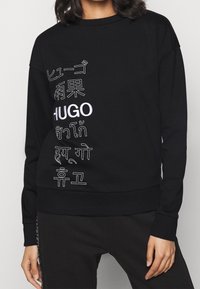 Sudadera negra con un diseño de texto audaz en varios idiomas. Cuello redondo, mangas largas y puños y dobladillo acanalados. Tela suave y texturizada.