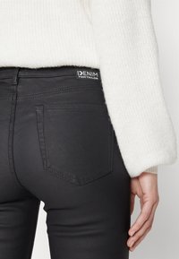 Acercamiento de una persona que lleva jeans de mezclilla negros con un suéter de punto blanco, mostrando el bolsillo trasero y la etiqueta de la marca "Denim Tom Tailor."