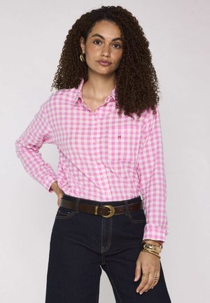 Donna con capelli ricci che indossa una camicia a quadri rosa e bianca, jeans scuri, cintura marrone e gioielli d'oro, posa con una mano sul fianco.