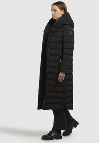 Vrouw in een lange zwart gewatteerde jas met grote kraag en capuchon, zwarte broek en zwarte enkellaarzen, die zijwaarts staat op een witte achtergrond.