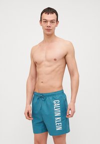 Tamsiai žalios maudymosi šortai su elastingu juosmeniu ir raišteliu. Kairėje pusėje yra balta "CALVIN KLEIN" ženklas. Lengvas audinys.