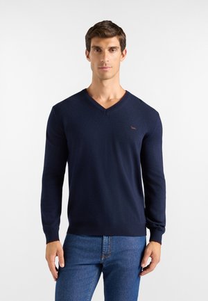 Uomo che indossa un maglione blu navy a scollo a V e jeans blu, in piedi davanti a uno sfondo bianco semplice.