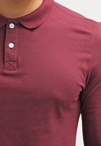 Burgunderfarbene Poloshirt aus strukturiertem Baumwollstoff. Verfügt über einen klassischen Kragen, eine Dreiknopfleiste und gestickte Akzente am Kragen.