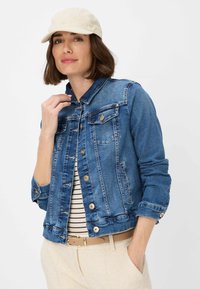 Giubbotto di denim di blu medio con due tasche sul petto, bottoni dorati e colletto classico. Indossato sopra una maglia a righe, abbinato a pantaloni chiari.
