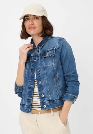 Giubbotto di denim di blu medio con due tasche sul petto, bottoni dorati e colletto classico. Indossato sopra una maglia a righe, abbinato a pantaloni chiari.