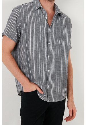 Mann trägt ein kurzärmliges, graues Hemd mit weißen vertikalen Streifen und schwarze Hose, mit einer Hand in der Tasche.