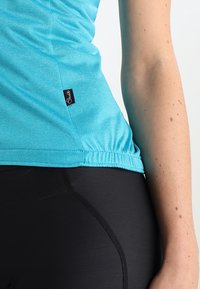 Top de tirantes ajustado de color azul claro con dobladillo elástico fruncido, combinado con leggings negros de alta cintura. Etiquetas visibles en el lado de la camiseta.