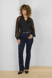 Juoda satino bluzonė su nėrinių rankovėmis, derinama su tamsiai mėlynomis platėjančiomis džinsomis ir juodais aukštakulniais bateliais su smailiu nosiu. Fonas neutralus.