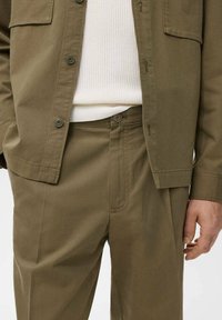 Camisa abotonada verde oliva con bolsillo en el pecho, combinada con pantalones a juego. La tela es suave, con costuras visibles y una capa base blanca texturizada y ribeteada.