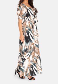 Kobieta w długiej sukience o abstrakcyjnym wzorze, z bufiastymi rękawami i czarnych sandałach na wysokim obcasie, stojąca w profilu na białym tle.