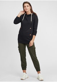 Felpa con cappuccio nera con cordini color crema e dettagli triangolari marroni, abbinata a pantaloni cargo joggers verde oliva e sneakers bianche.