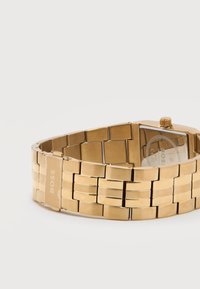 Πολύχρυση μεταλλική μπάντα ρολογιού χειρός με αγκράφα χαραγμένη με τη λέξη "BOSS", εμφανιζόμενη από την πίσω πλευρά και ελαφρώς τυλιγμένη σε ουδέτερο φόντο.