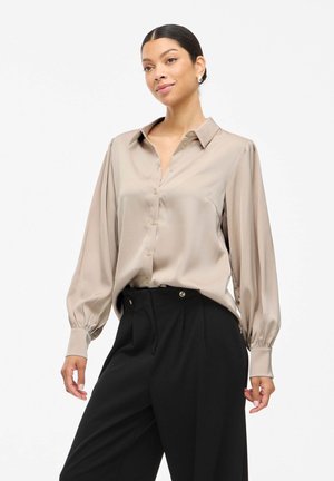 Kvinne iført beige satengbluse med lange puffede ermer og svarte vide bukser mot en hvit bakgrunn.