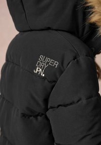 Veste matelassée noire avec capuche, logo brodé "SUPER DRY JPN." sur la poitrine, bordure en fourrure visible au bord de la capuche.