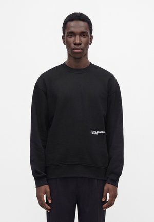 Jonge man staand tegen een witte achtergrond, draag een zwarte lange mouwen trui met de tekst "Karl Lagerfeld Jeans" aan de voorkant.