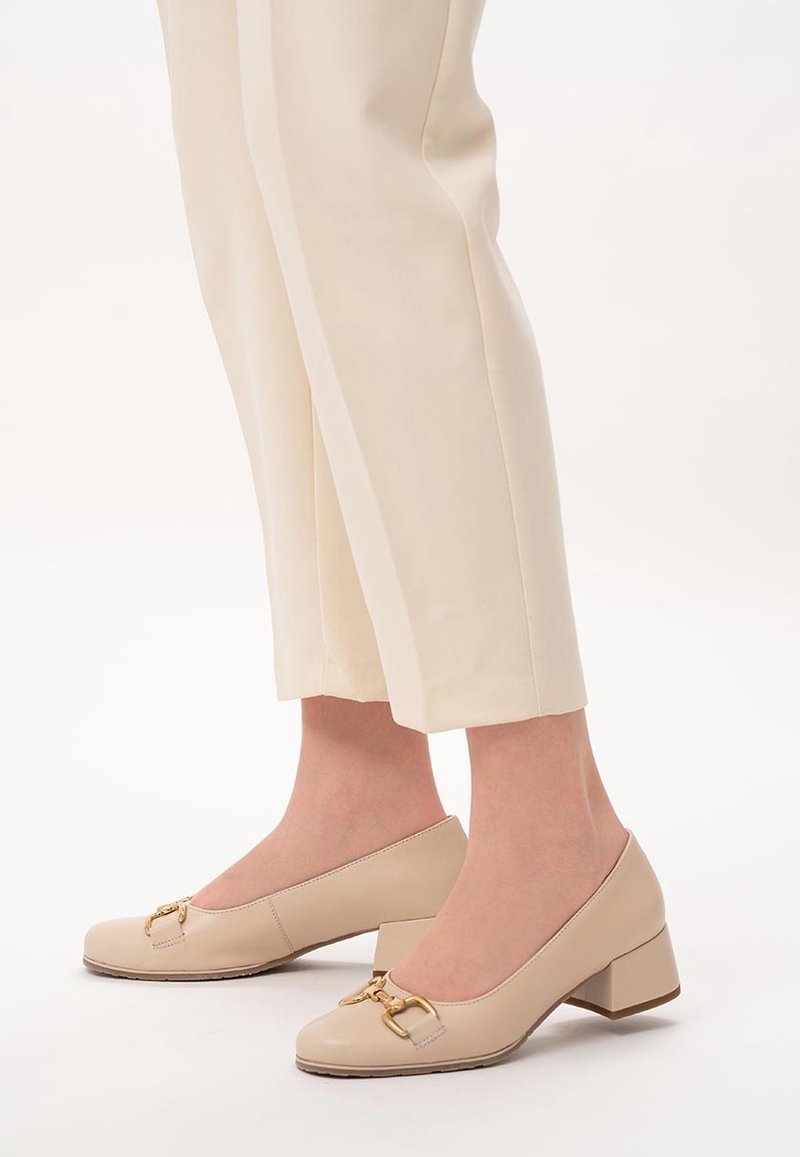 Chaussures pour femmes beige à talons bas avec détail boucle dorée, portées avec un pantalon court crème sur fond blanc.