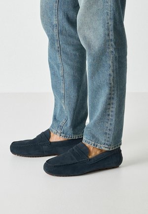 Benedenbenen in lichtblauwe spijkerbroek en donkerblauwe suède loafers met rubberen zolen, staand op een lichte vloer tegen een effen muur.