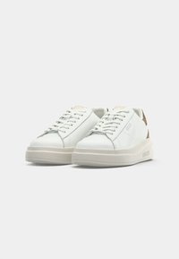 Sneakers bianchi a bassa punta con suole spesse beige, design con lacci e dettagli in marrone sul tallone, posizionati uno accanto all'altro su uno sfondo bianco.