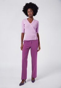 Lilas stickad omslagsblus med korta ärmar, vågformad halsringning och knappdetalj, i kombination med rak benlängd lila byxor och klackskor.