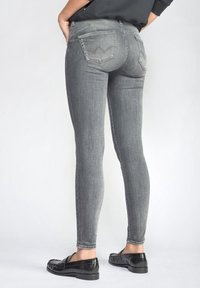 Osoba v přiléhavých šedých džínech a černých mokasínech, stojící proti jednobarevnému světlému pozadí, s rukama opřenýma o boky.