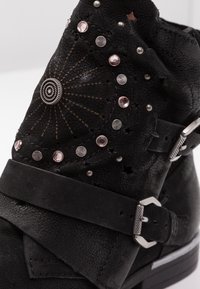 Black ankle boot made of textured leather with metallic studs, a decorative medallion, and a strap with a silver buckle.