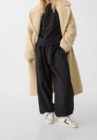 Beige schapenleren jas over een zwart sweatshirt en brede zwarte broeken. Witte sneakers met zwarte accenten. Zachte texturen en een ontspannen pasvorm.