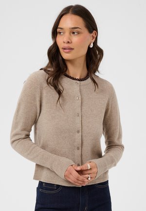 Kvinde vendt mod venstre iført beige cardigan med knapper, mørke jeans, perleøreringe, mørkt perlehalskæde og ringe mod en ensfarvet baggrund.