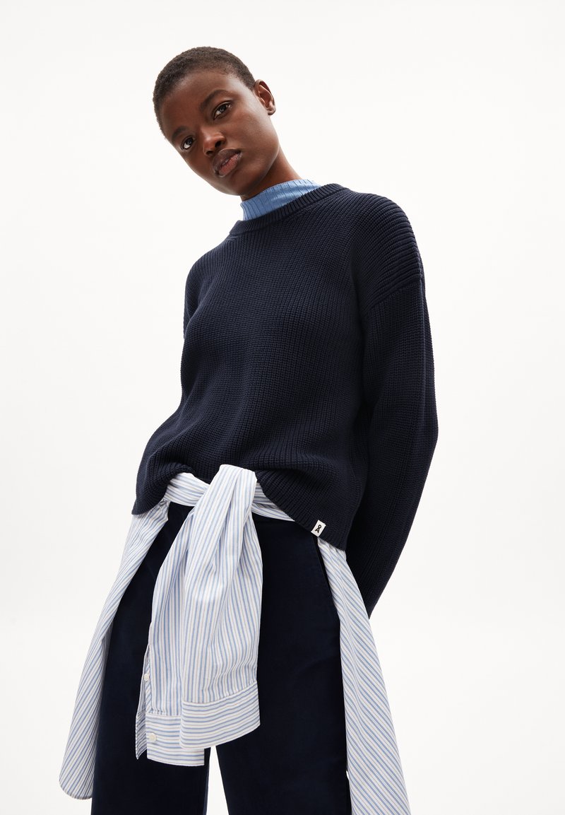 Marineblauer Strickpullover mit gerippter Textur, ergänzt durch ein hellblau gestreiftes Hemd, das um die Taille gebunden ist. Auffälliges Etikettendetail auf dem Pullover.