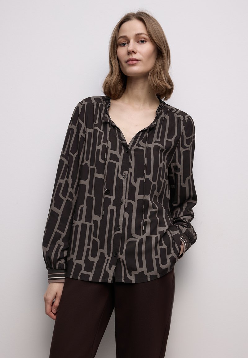 Blouse à motif sombre avec un design géométrique en noir et gris, avec un col en V, des manches longues et une fermeture par boutons.