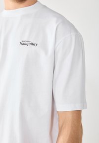 Hvit bomulls T-skjorte med korte ermer, med teksten "Soar into Tranquility" i svart på brystet. Enkel og udecorert design.