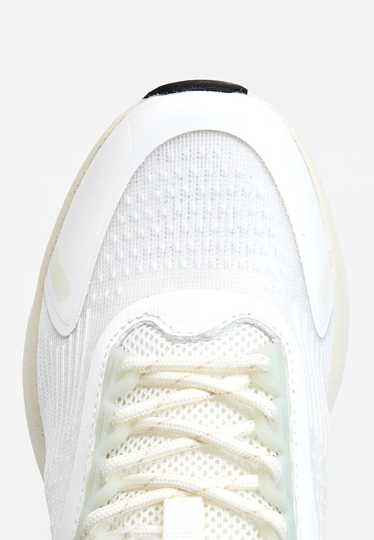 Zapatilla deportiva blanca con parte superior de malla texturizada, acentos en color crema y cordones planos; cuenta con una suela de goma suave y una etiqueta negra en el talón.