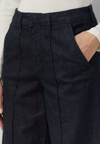 Dunkelblaue Jeans mit hoher Taille, geradem Schnitt, Seitentaschen und sichtbarer Naht entlang der Nähte. Glatte Textur.