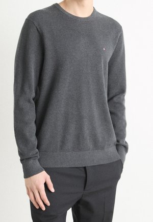 Muž v tmavě šedém pleteném svetru s dlouhým rukávem a černých kalhotách, stojící s jednou rukou v kapse na jednobarevném pozadí.