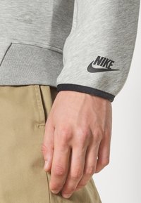 Acercamiento de una mano descansando junto al bolsillo de un pantalón beige, con un sudadero gris de Nike que tiene el logotipo negro en el puño de la manga.