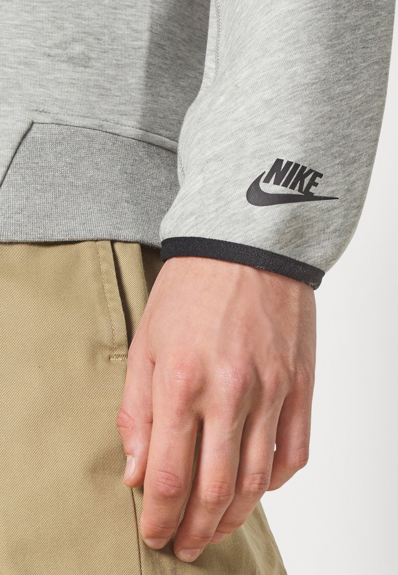 Gros plan sur une main reposant près de la poche d'un pantalon beige, portant un sweat-shirt Nike gris avec un logo noir sur le poignet.