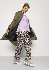 Olive green quilted long coat, lavender top, and floral patterned pants. Gray canvas sneakers. Smooth fabrics with vibrant colors and intricate designs.
