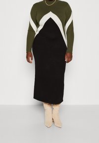 Abito in maglia con un top verde oliva decorato da motivi a zig zag bianchi e una gonna nera, completato da stivali beige con punta affusolata.