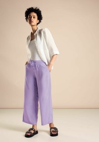 Luźne, lawendowe spodnie z elastycznym paskiem w talii, zestawione z białą koszulą na guziki i czarnymi sandałami z metalowymi akcentami.