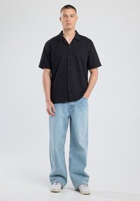 Black short-sleeve button-up shirt with a collar, paired with light blue wide-leg jeans and white sneakers. Smooth fabric, relaxed fit.