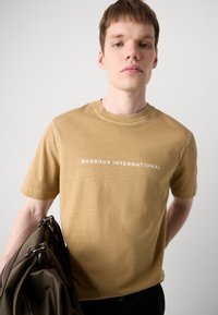  Helepruun puuvillane T-särk lühikeste varrukatega ja ümar kaelus, millel on rindades valge tekst "BARBOUR INTERNATIONAL".