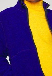 Polarowa kurtka w głębokim niebieskim kolorze z kontrastowym czarnym zamkiem błyskawicznym; posiada jasnonośny golf w kolorze żółtym oraz kieszeń z przodu.